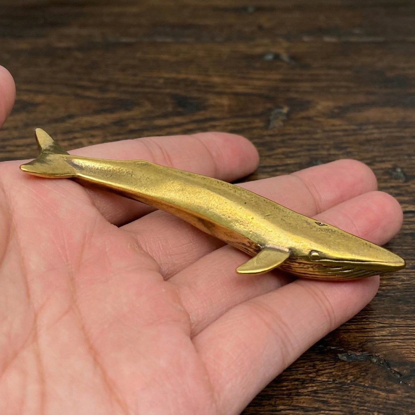 The Ancient Navigator miniature brass whale held in hand to show scale, with detailed flukes and textured patina, from the WILLAGE Tiny Objects collection.