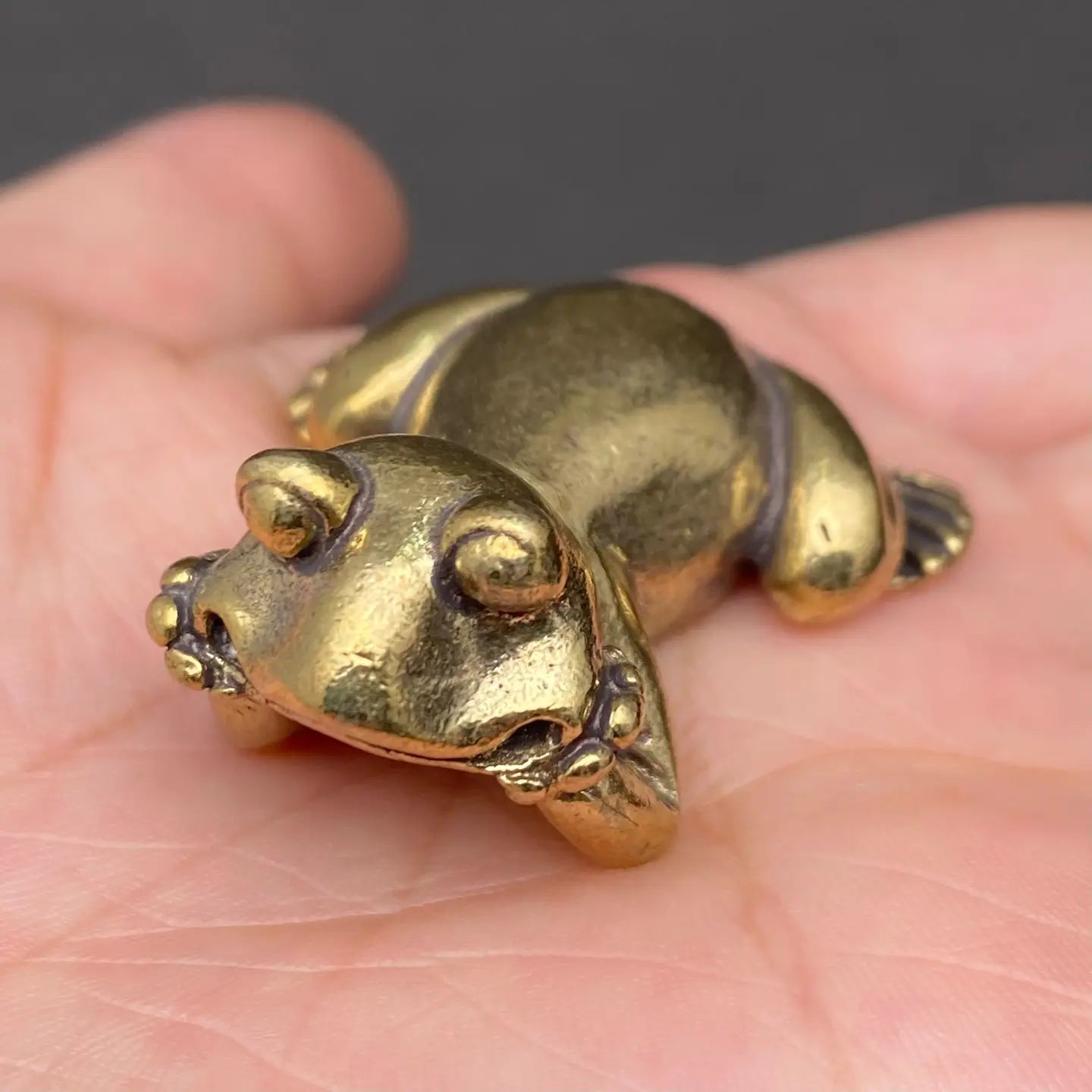 The Jovial Companion miniature brass frog held in hand to show scale, with playful reclining pose and golden patina