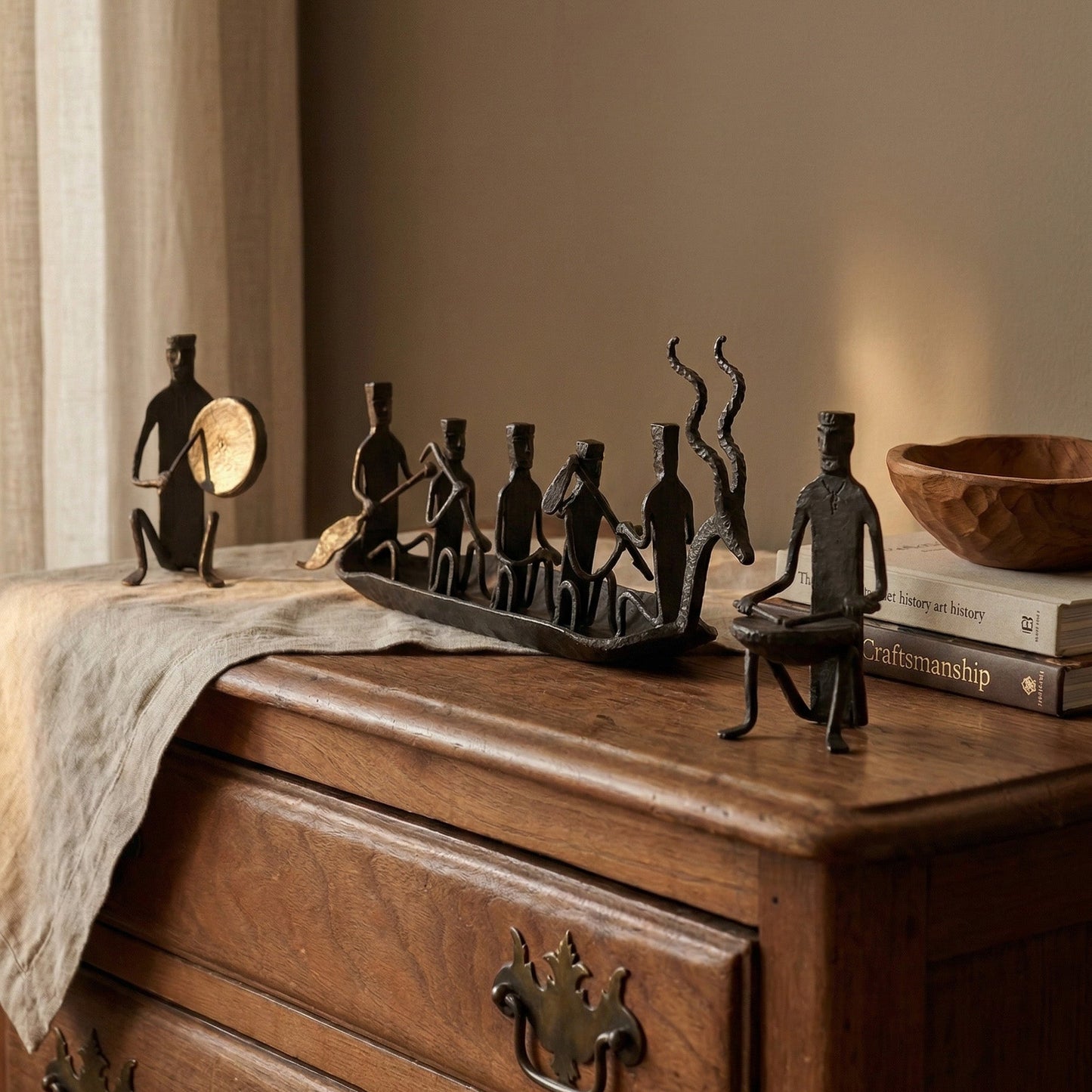 The Music Boat Bastar wrought iron sculpture displayed on an antique wooden dresser with linen runner, craftsmanship books, and companion gong-playing and drummer figures, hand-forged in Chhattisgarh India, from WILLAGE