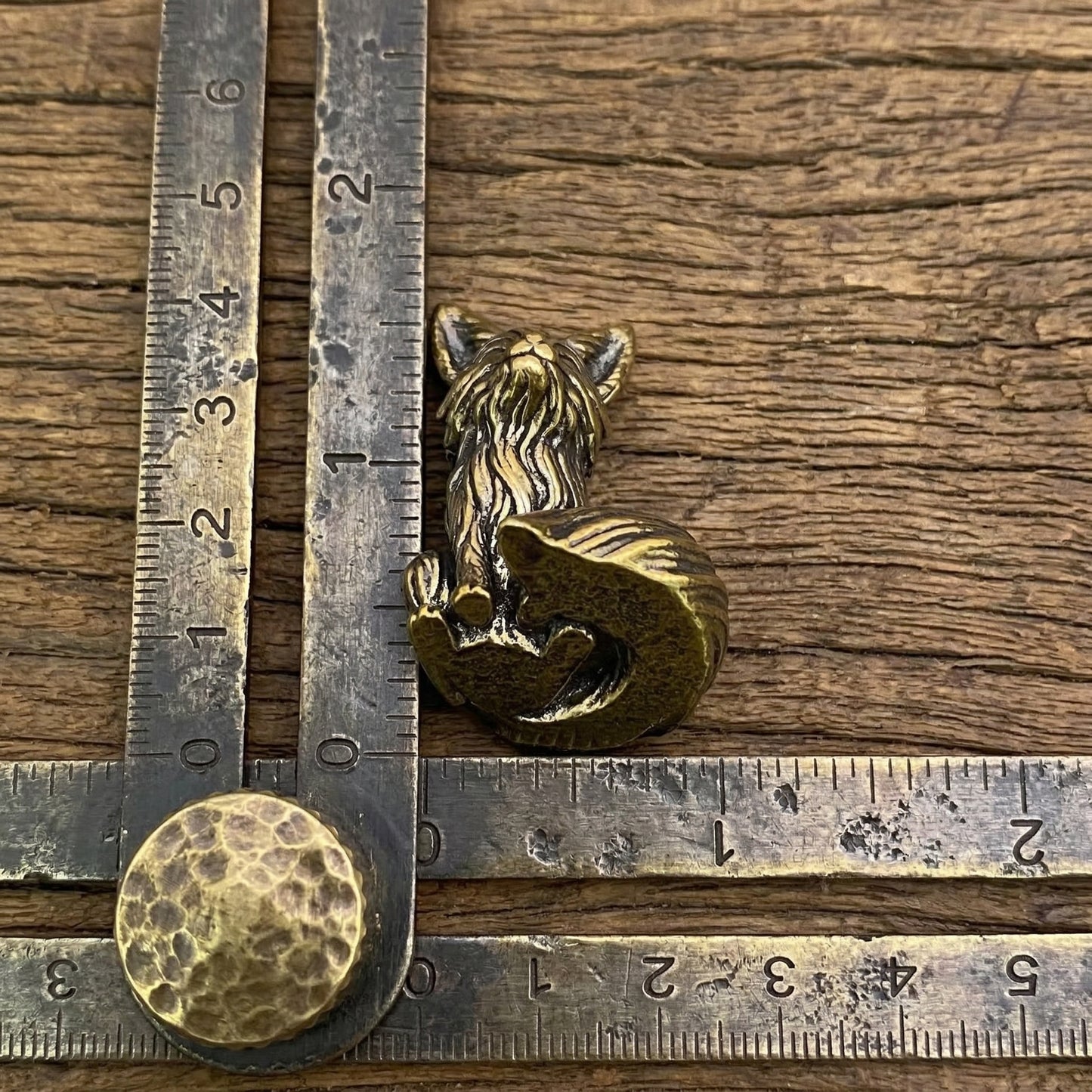 The Playful Kit miniature brass baby fox shown with ruler for scale, approximately 40 mm tall, with detailed fur texture and aged patina on a wooden surface