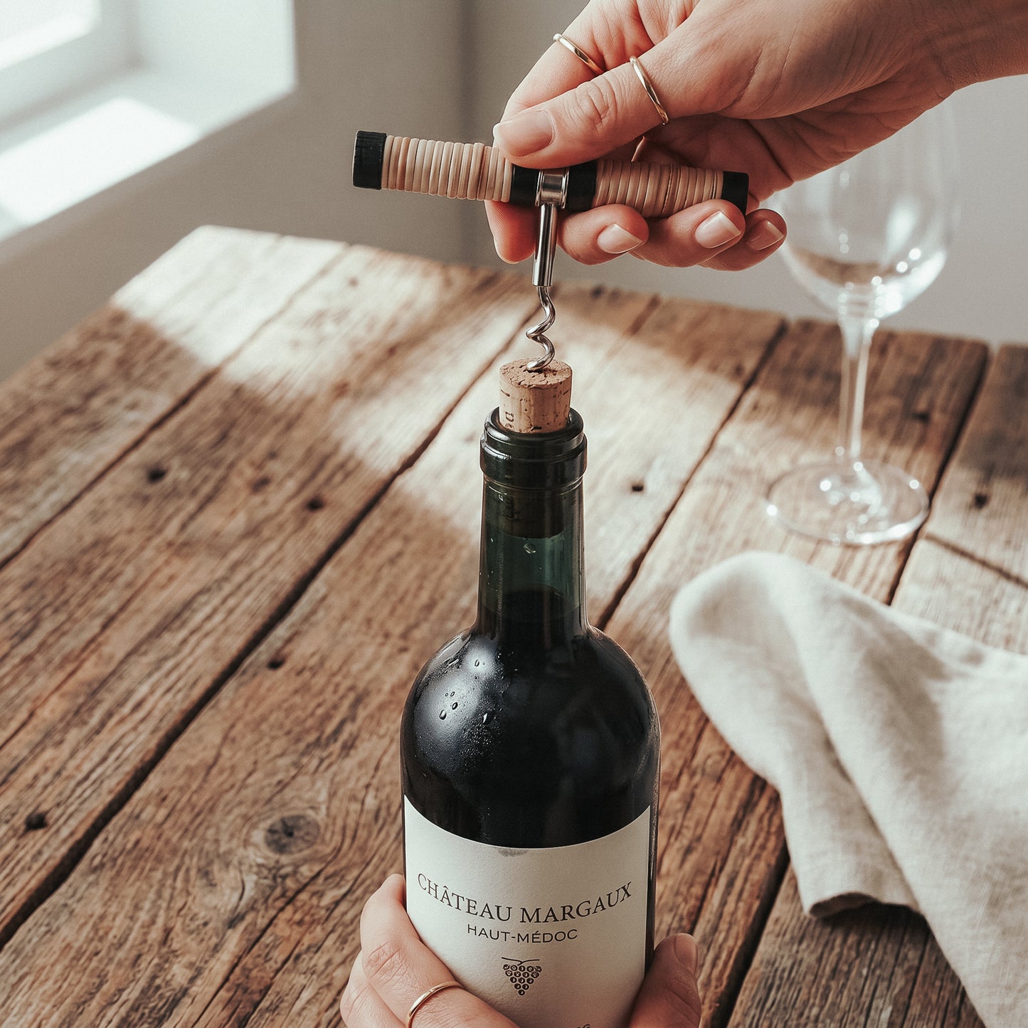 The Rattan Corkscrew being used to pull a cork from a wine bottle on a rustic wood table, showing its compact pull-style size
