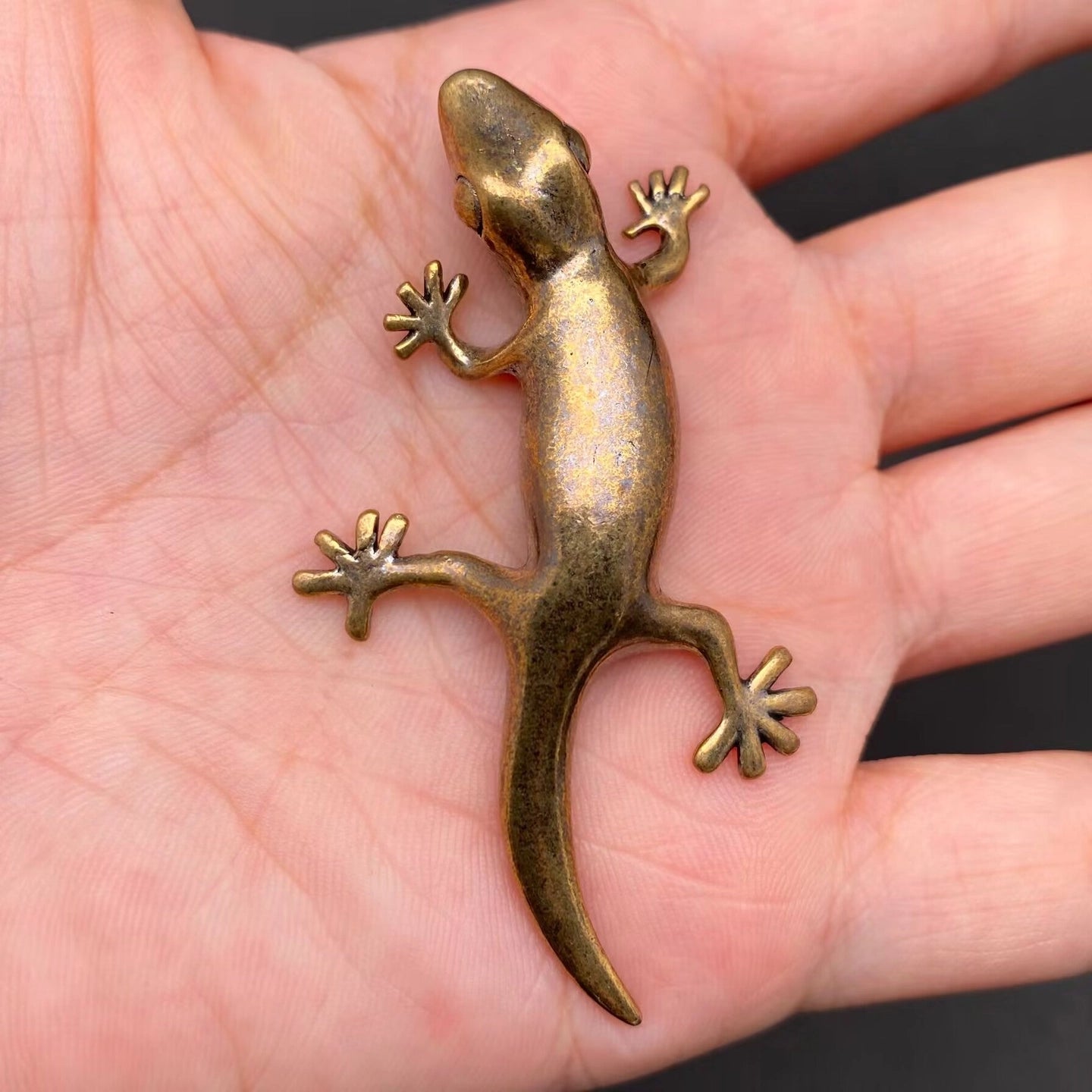 The Sunlit Wanderer miniature brass gecko in a crawling pose with splayed limbs and curved tail, showing warm aged patina on a wooden surface, from the WILLAGE Tiny Objects collection