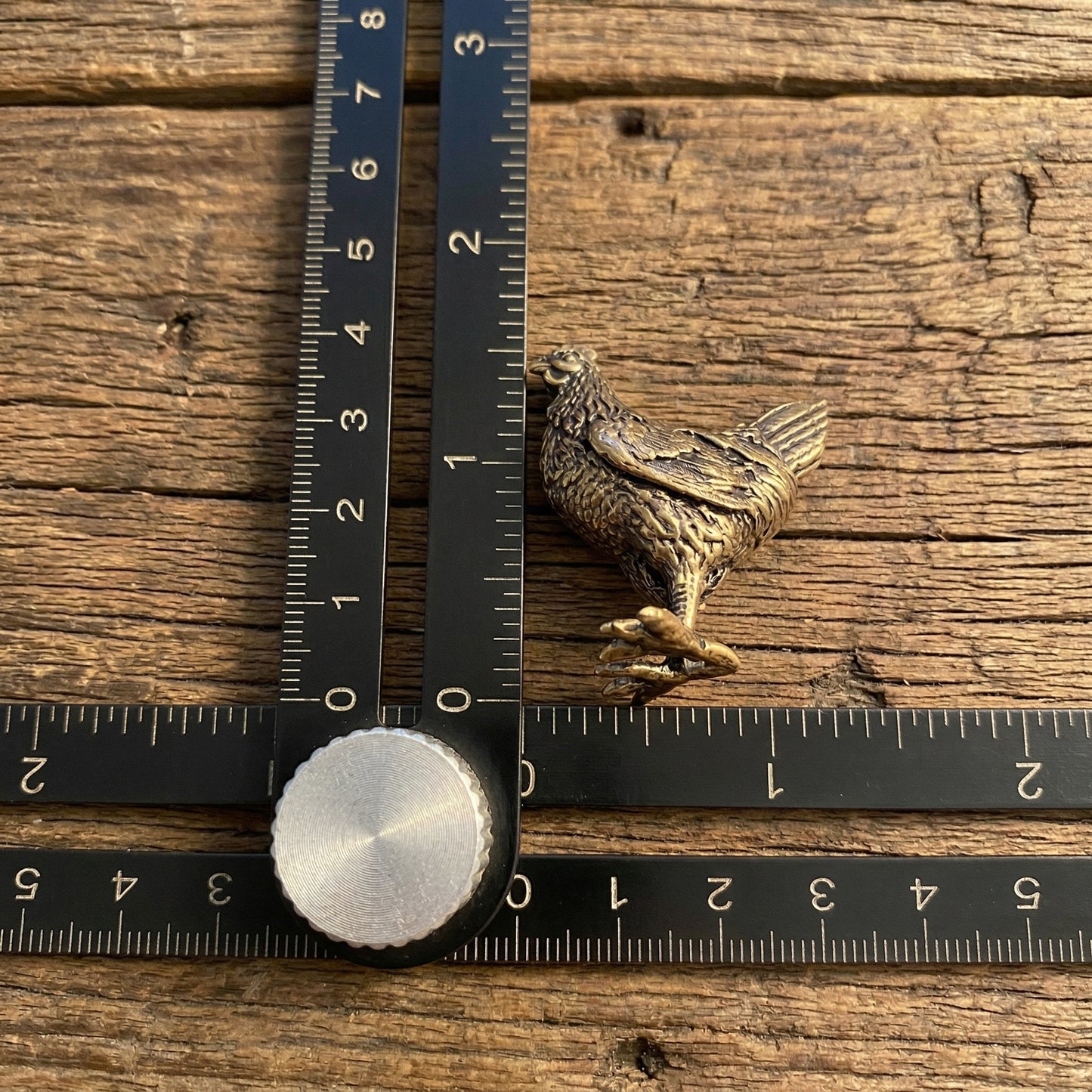 The Vigilant Nurturer miniature brass hen shown with ruler for scale, approximately 39 mm tall, with detailed feathers and aged patina on a wooden surface