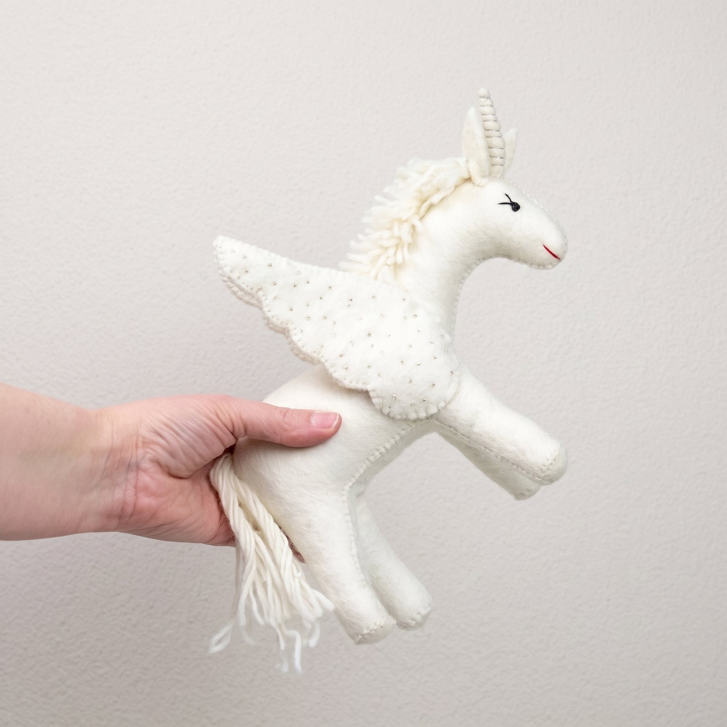 White felted unicorn held in a hand, showing its beaded wings and flowing wool tail