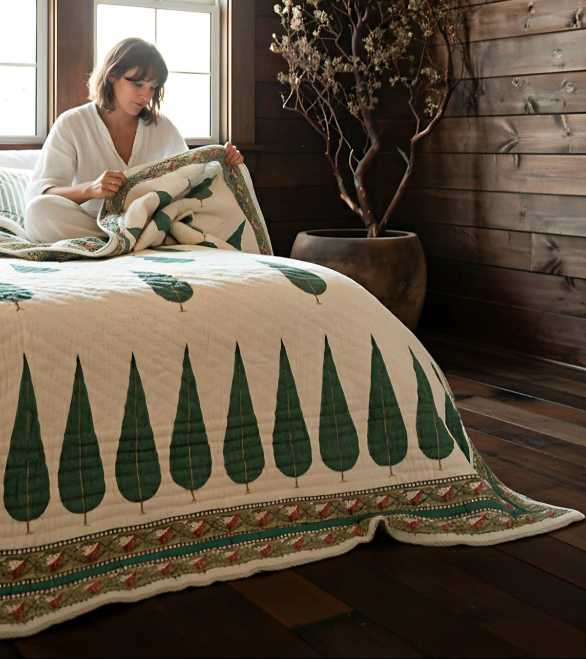 Woman sitting on a bed with a patterned blanket in a room with wooden walls and a large window.