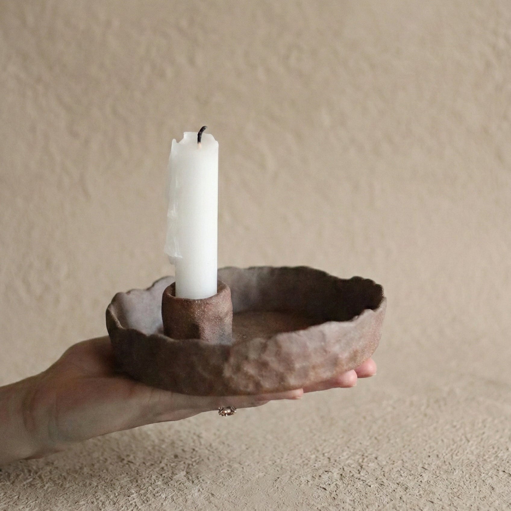 Hand holding a rustic  candle holder with a white candle against a beige background
