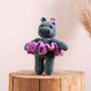 A crocheted hippo ballerina in a tutu, primarily in blue and purple colors, perched on a wooden stump.