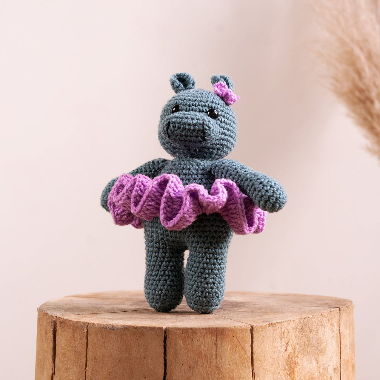 A crocheted hippo ballerina in a tutu, primarily in blue and purple colors, perched on a wooden stump.