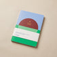 Reading journal with a red sun over green field and blue sky cover, gold-foil title, rounded corners, on a beige surface