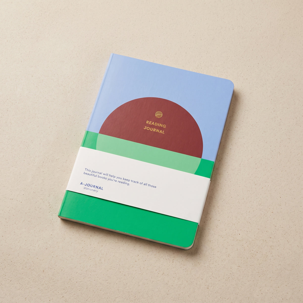 Reading journal with a red sun over green field and blue sky cover, gold-foil title, rounded corners, on a beige surface