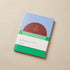 Reading journal with a red sun over green field and blue sky cover, gold-foil title, rounded corners, on a beige surface