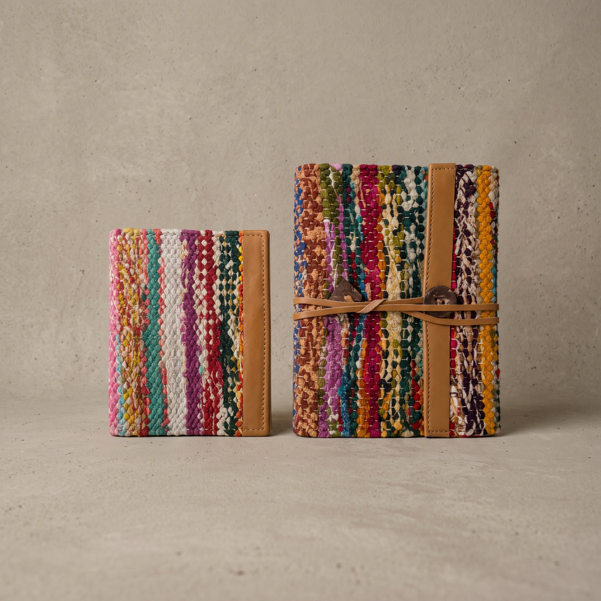 Two closed handmade journals with a multicolor cover woven from recycled sari cloth, tan leather strap with button, and a wrap tie ending in a heart-shaped charm