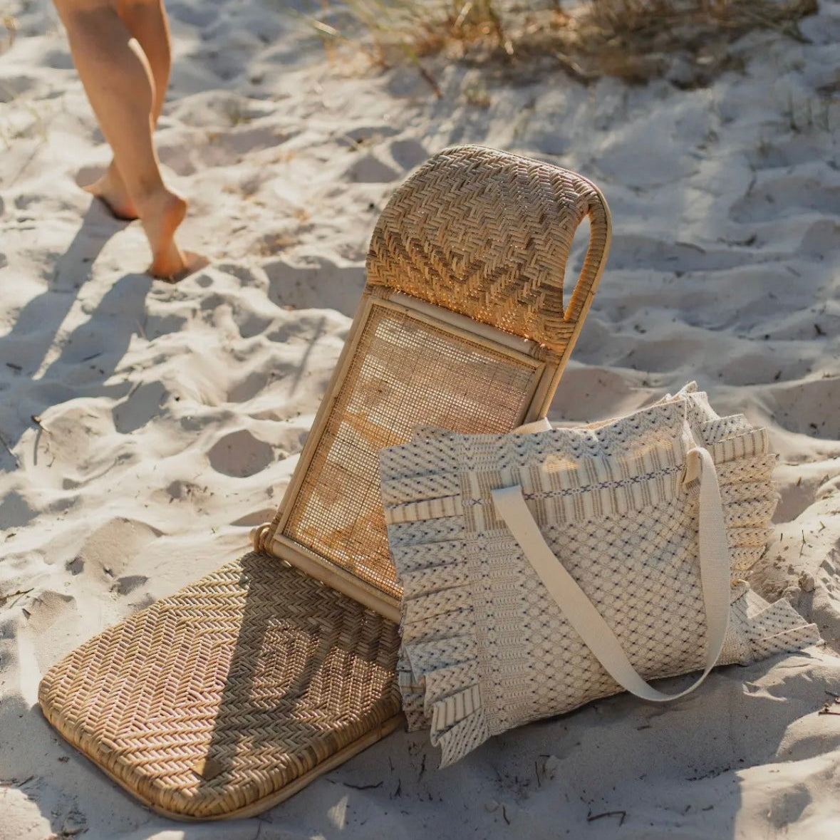 A straw chair and a matching straw bag on a sandy beach with a person walking in the background.