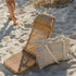 A straw chair and a matching straw bag on a sandy beach with a person walking in the background.