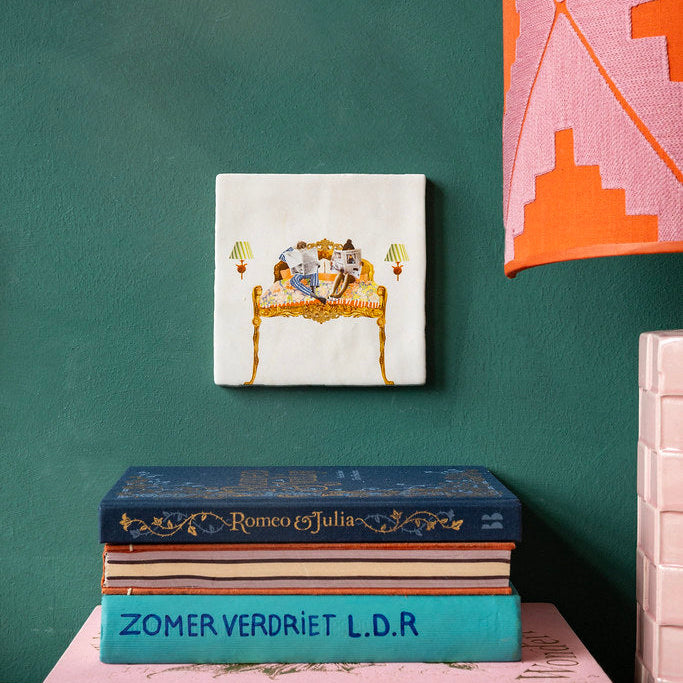 Best Beginning of the Day Storytile propped on a shelf with Dutch books on a teal wall, showing illustrated gilded bed scene, hand-baked ceramic by Marga van Oers in the Netherlands, from WILLAGE