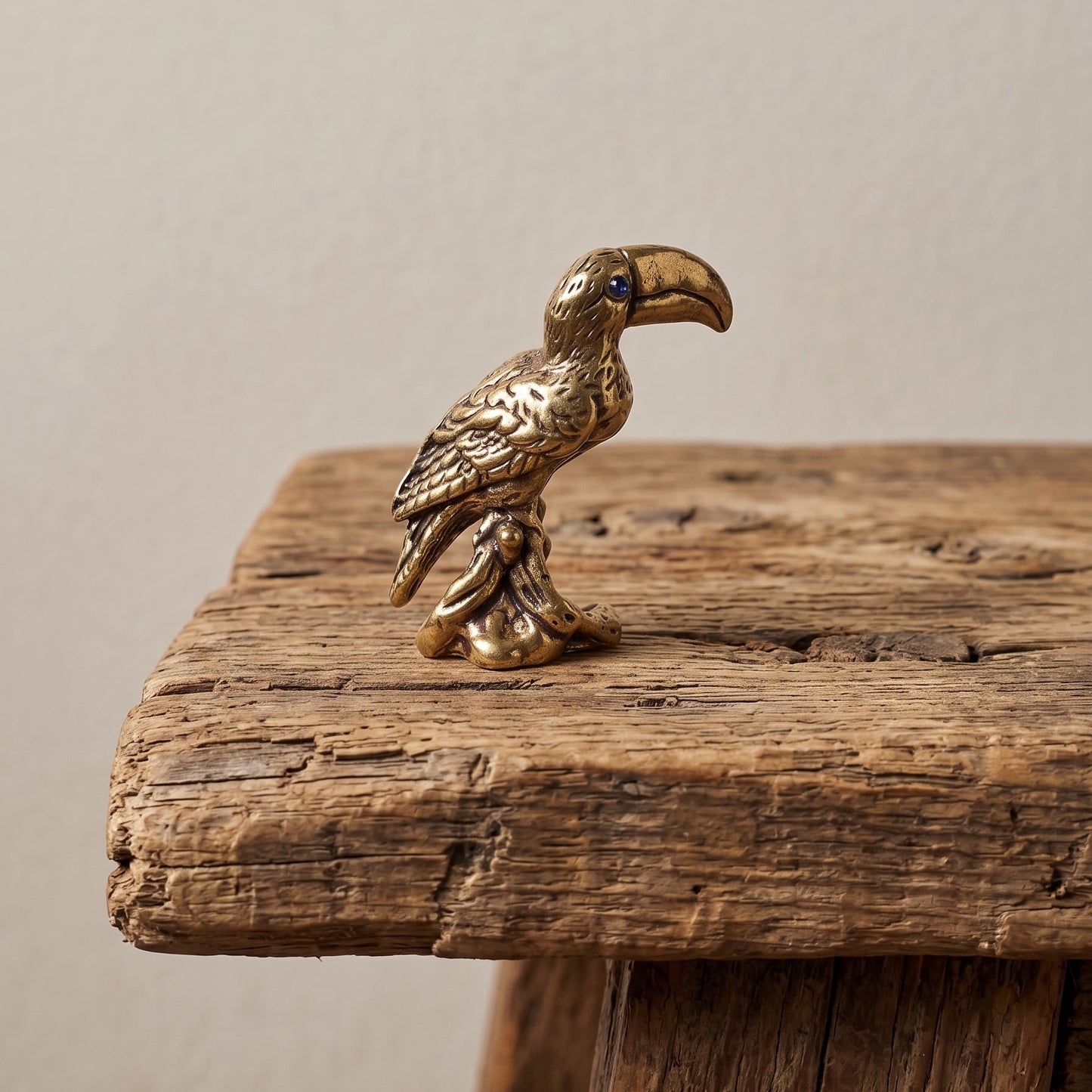 Small solid brass toucan figurine perched on a branch with a raised bill, fine feather detail and a blue stone eye, on weathered wood