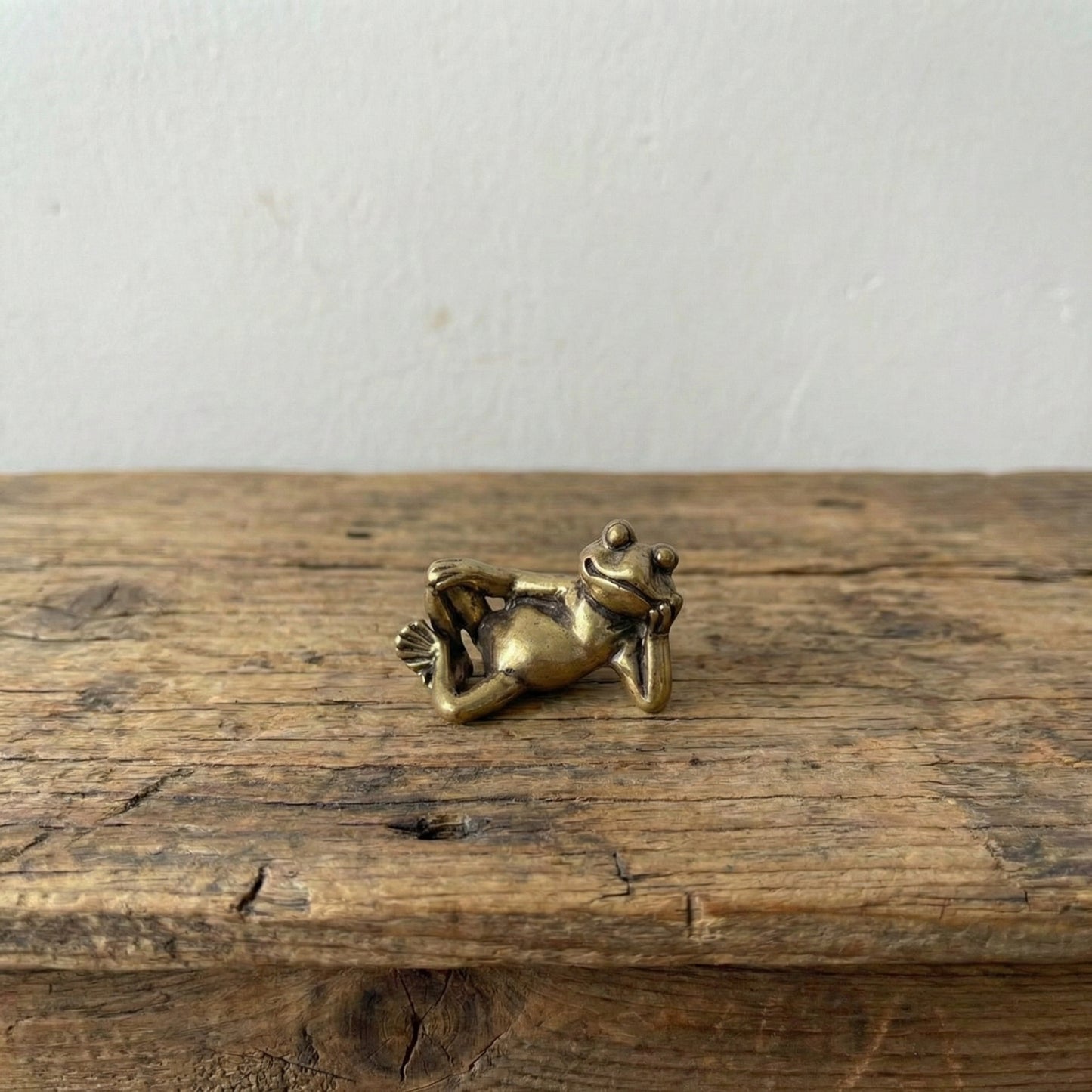 The Content Spirit miniature brass frog reclining on its side with a smirk, showing warm aged patina on a wooden surface, from the WILLAGE Tiny Objects collection