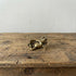 The Content Spirit miniature brass frog reclining on its side with a smirk, showing warm aged patina on a wooden surface, from the WILLAGE Tiny Objects collection