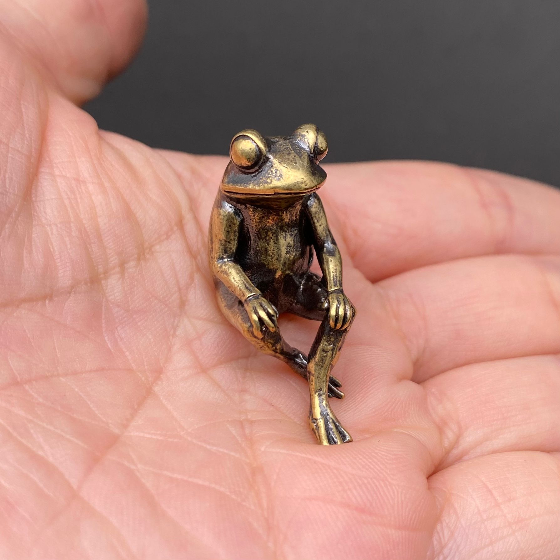 The Daydreamer brass frog held in a hand to show its small size, solid brass with warm patina