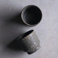Short unglazed Iga ware stoneware cup in dark, pebbled clay with a tapered foot, on a white background