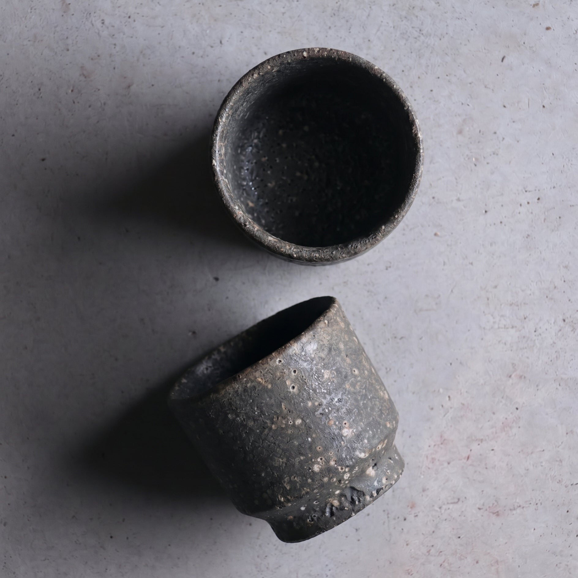 Short unglazed Iga ware stoneware cup in dark, pebbled clay with a tapered foot, on a white background