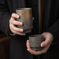 Two unglazed Iga ware stoneware cups — the taller large size and the shorter small size — held in two hands against dark linen clothing, showing the rough pebbled clay and warm bronze shimmer