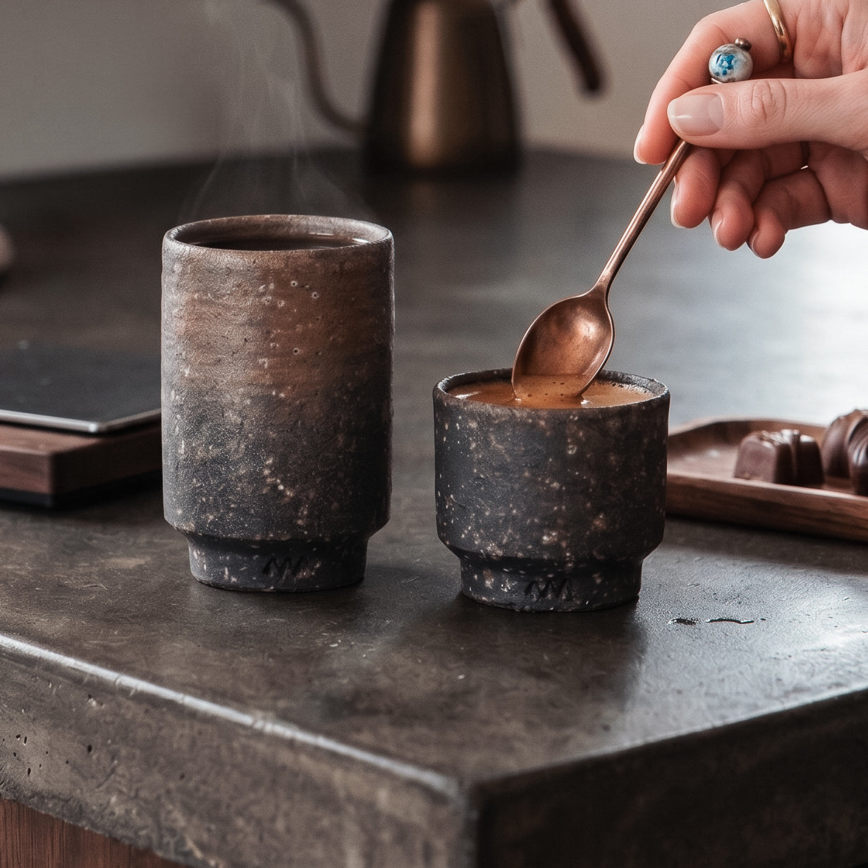 Two unglazed Iga ware stoneware cups in different sizes on a dark stone counter, one holding coffee with a copper spoon resting in it, rough pebbled clay and bronze shimmer catching the light
