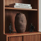 The Gathered Earth Vessel hand-built clay sculpture by Conrad and Dina displayed on a walnut shelf flanked by Bastar wrought iron figures and art books, from WILLAGE