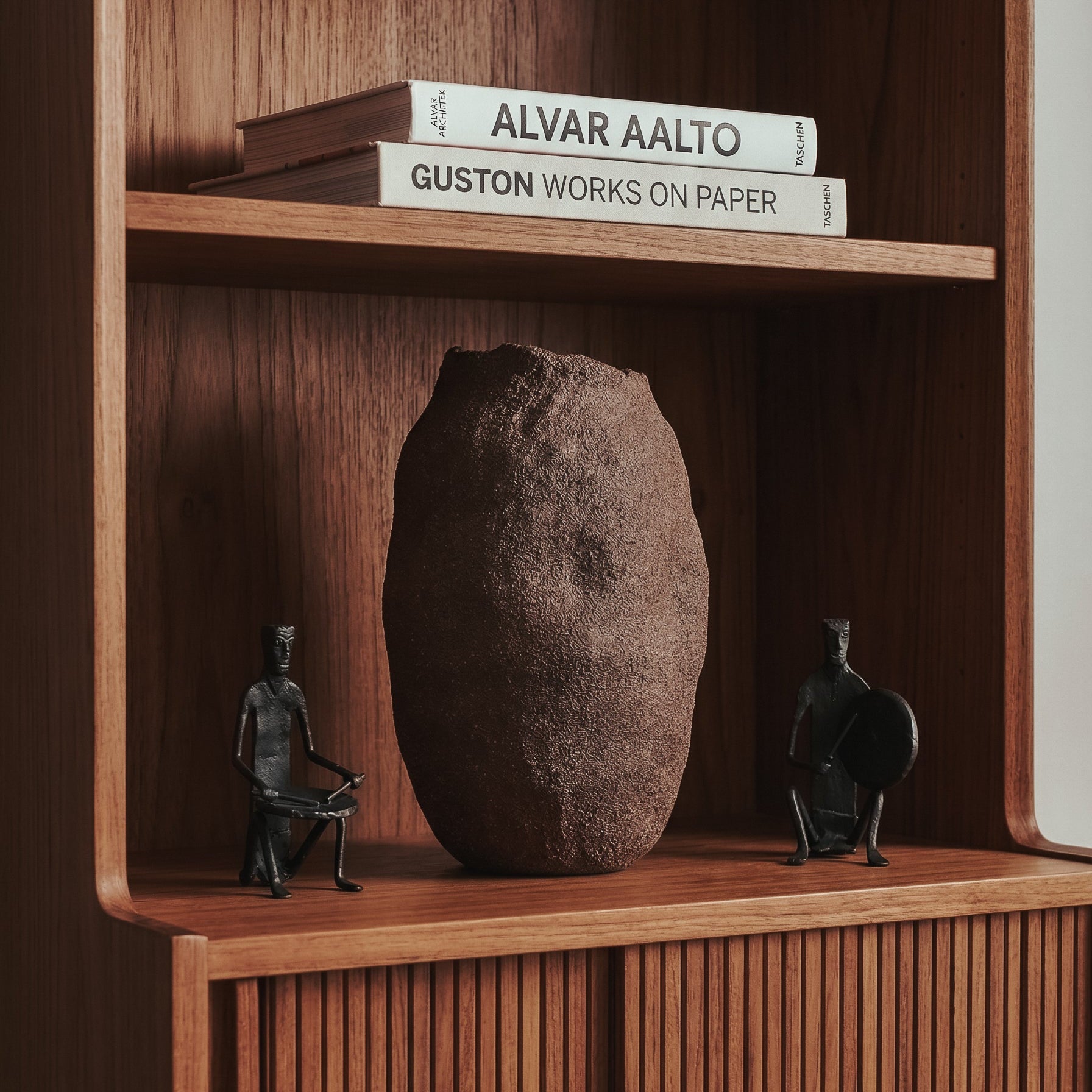 The Gathered Earth Vessel hand-built clay sculpture by Conrad and Dina displayed on a walnut shelf flanked by Bastar wrought iron figures and art books, from WILLAGE
