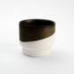 Porcelain cup half-dipped in dark bronze glaze over a raw white porcelain base, photographed on a pale gray background