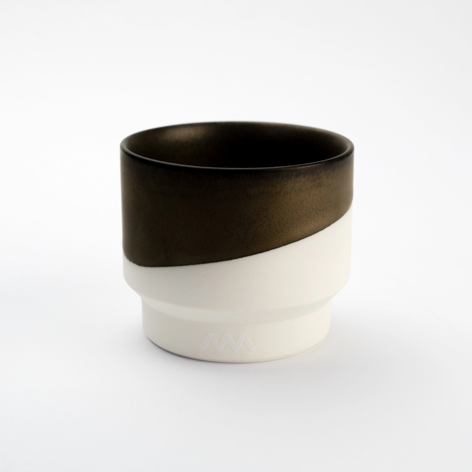 Porcelain cup half-dipped in dark bronze glaze over a raw white porcelain base, photographed on a pale gray background