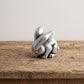 Small pewter rabbit netsuke with ears laid back, sitting on botanical-print paper