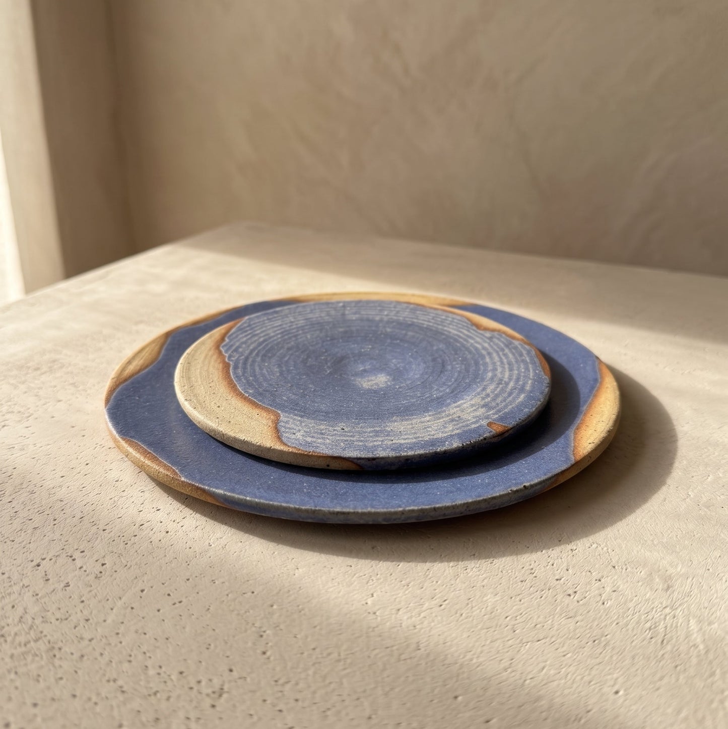 Large and small blue stoneware plates styled on white linen and a sunlit wooden floor beside a window, with a stoneware bowl resting on the small plate