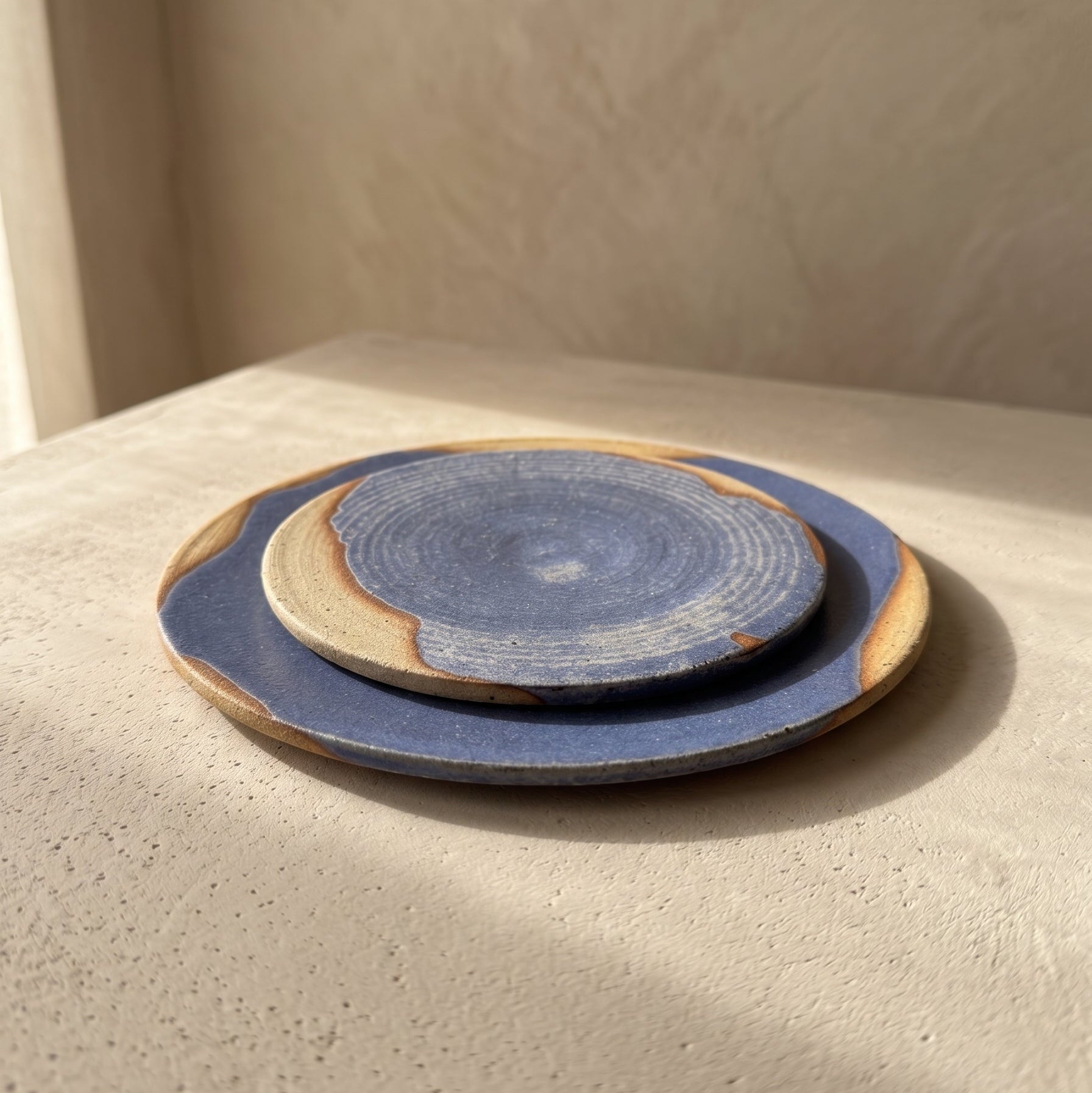 Large and small blue stoneware plates styled on white linen and a sunlit wooden floor beside a window, with a stoneware bowl resting on the small plate