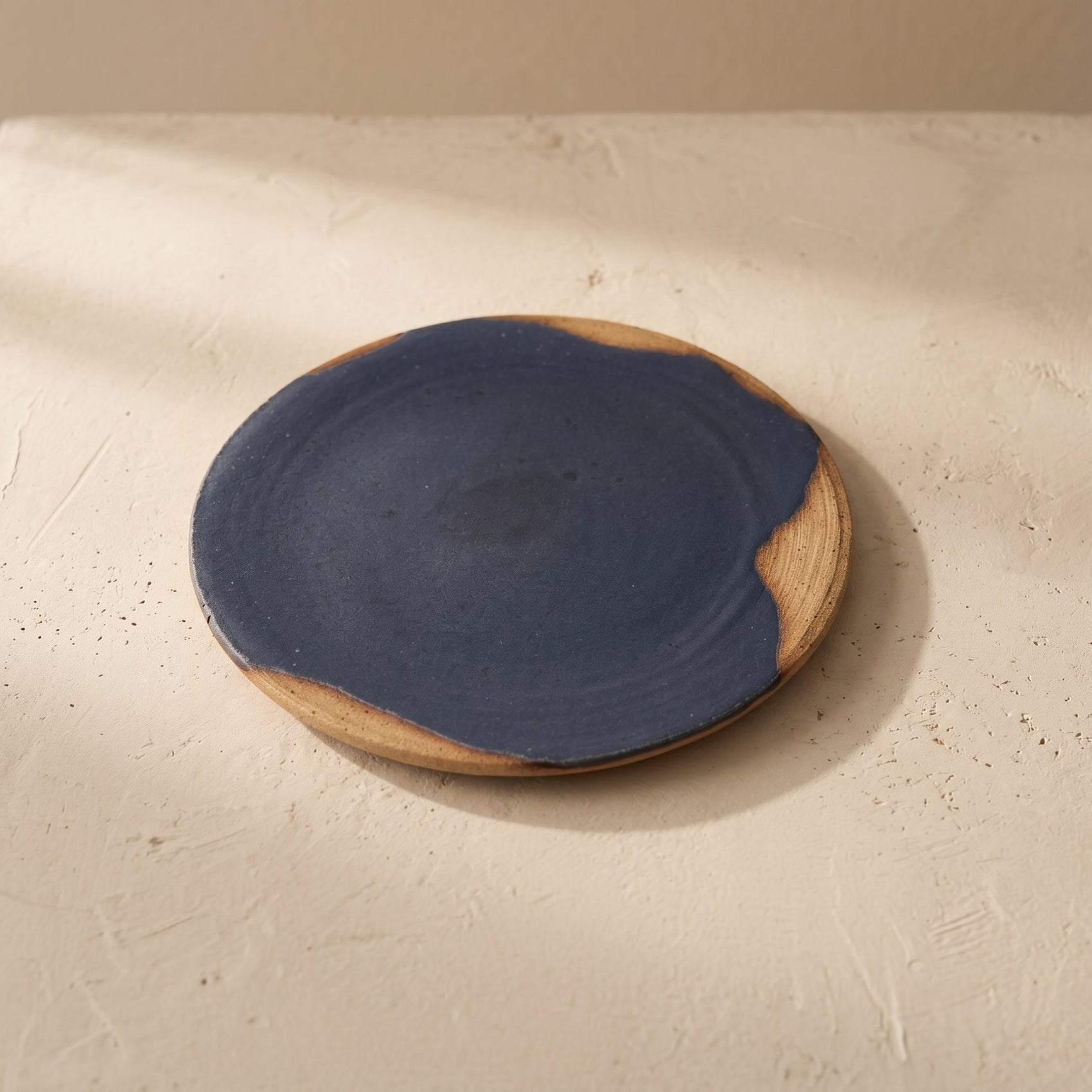 Large stoneware plate styled on white linen and a sunlit wooden floor beside a window, with a stoneware bowl resting on the small plate