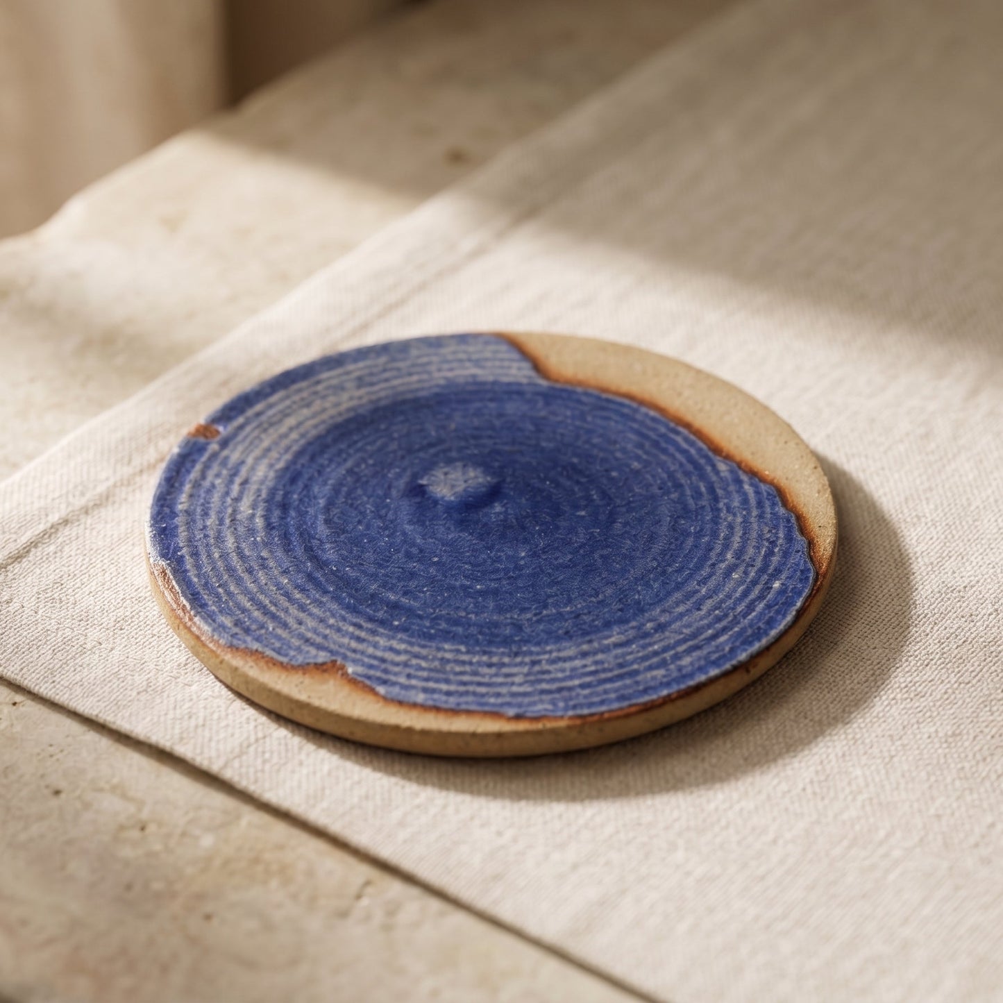 Small blue stoneware plate on natural linen, showing concentric wheel-thrown rings under the glaze and the bare clay edge