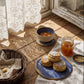 Blue stoneware plates holding scones, figs and jam on a sunlit wooden table beside a bread basket and a bowl of yoghurt and berries
