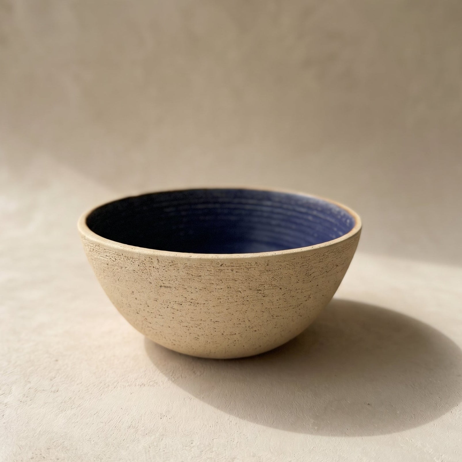 A handmade stoneware bowl with a deep blue glazed interior and a sand-textured unglazed exterior, lit from the side
