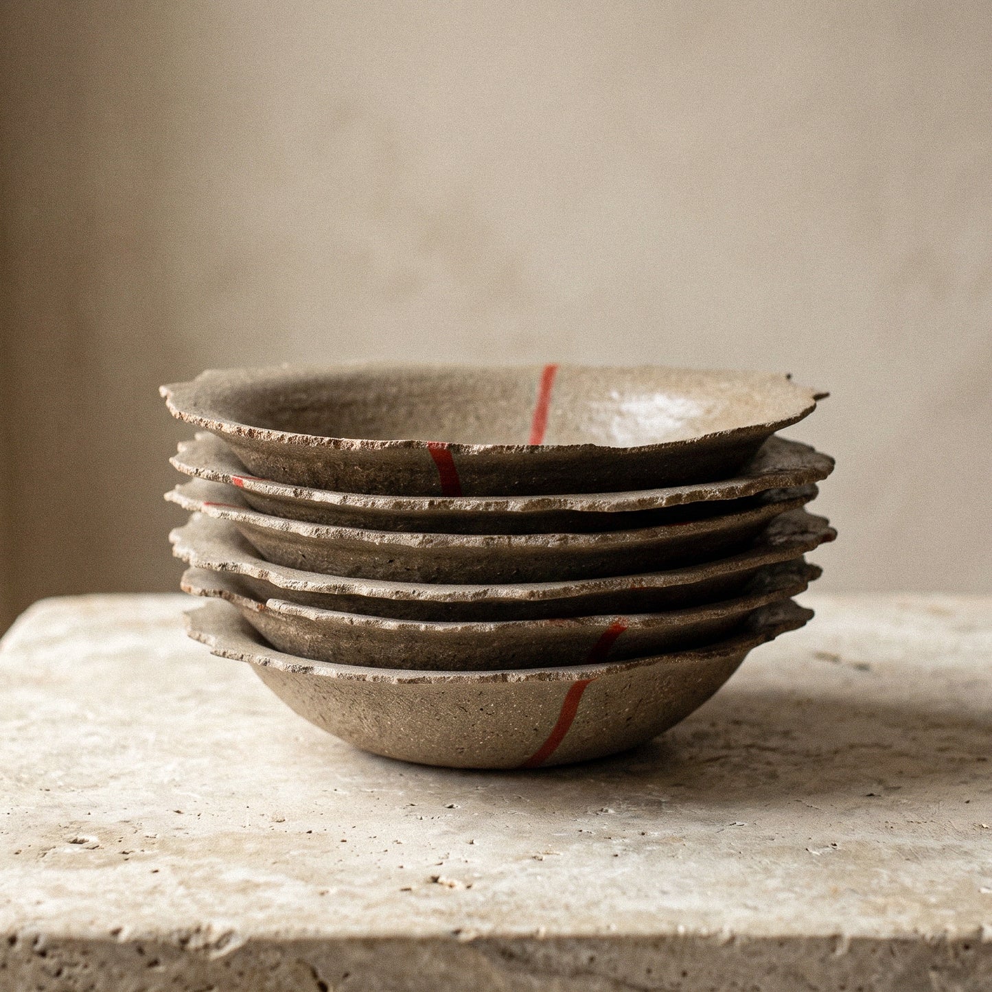 Six handmade stoneware plates stacked on a stone surface, each with an uneven organic edge and a red line, showing the variation between pieces