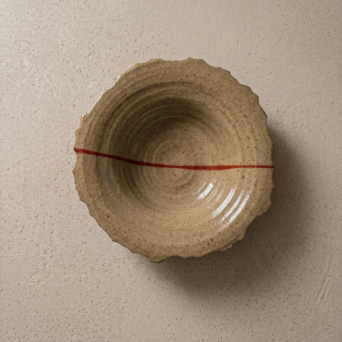 Handmade stoneware plate seen from above, sandy speckled clay with a torn organic edge, ridged wheel-thrown interior and a single red line running across it