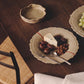 Handmade stoneware plate on a dark wood table holding blue cheese, dates and raisins, with cutlery and a second plate of grapes nearby