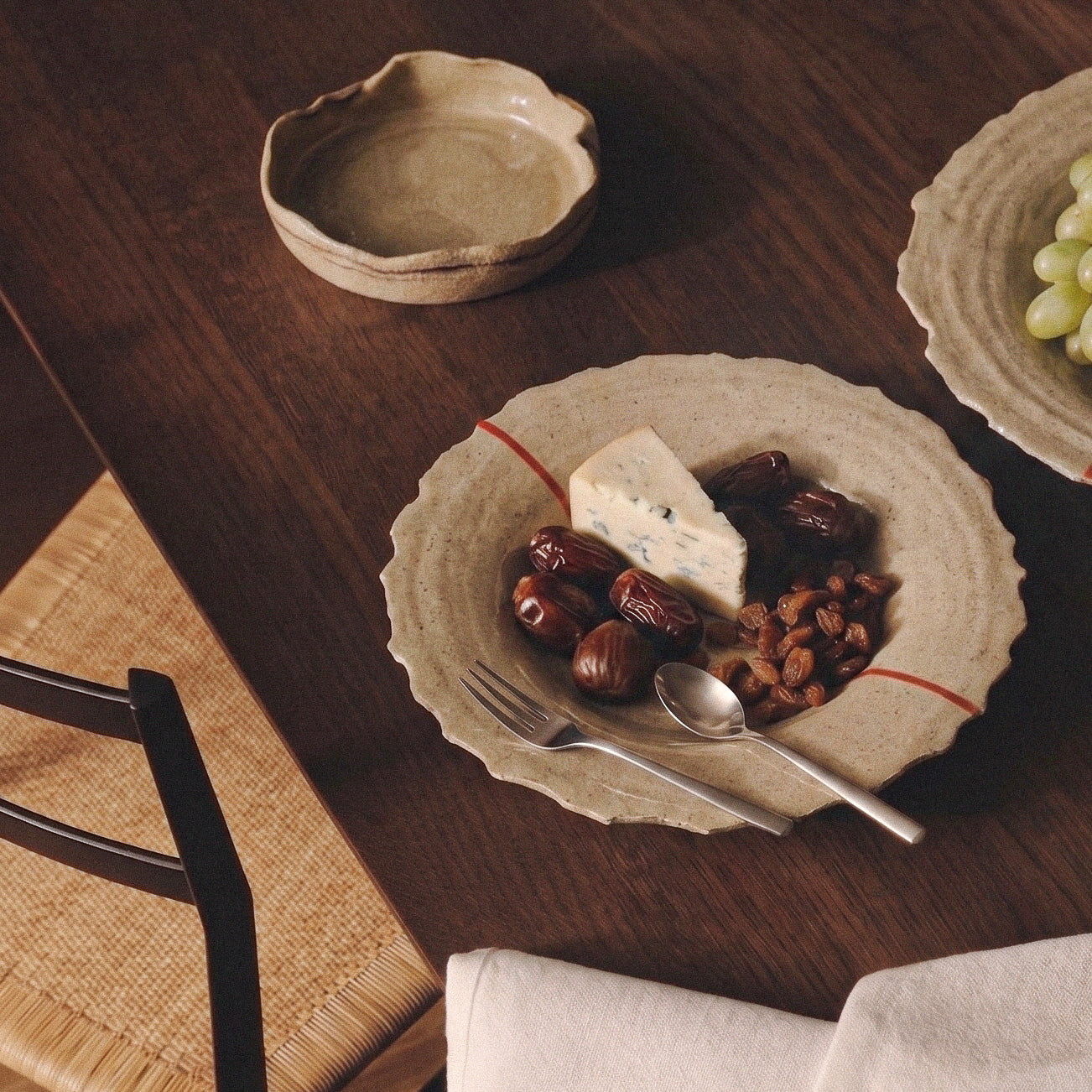 Handmade stoneware plate on a dark wood table holding blue cheese, dates and raisins, with cutlery and a second plate of grapes nearby
