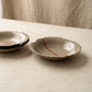 Two handmade stoneware plates on a pale surface, one angled to show the shallow curve, torn rim and glazed interior