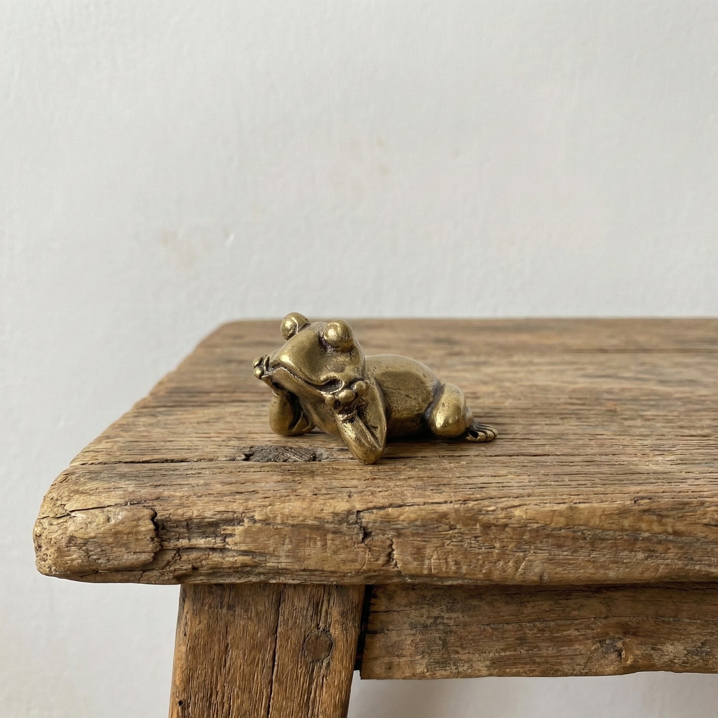 The Jovial Companion miniature brass frog reclining on its side with a grin, showing warm aged patina on a wooden surface, from the WILLAGE Tiny Objects collection