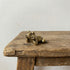 The Jovial Companion miniature brass frog reclining on its side with a grin, showing warm aged patina on a wooden surface, from the WILLAGE Tiny Objects collection