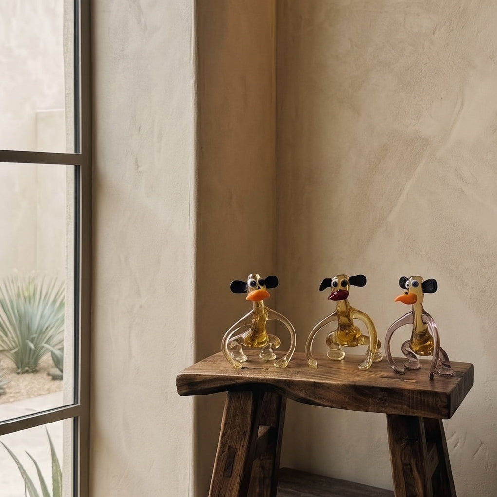 Three Lanky Ape glass monkey figurines displayed on a small wooden stool in a modern interior niche with books and a brass vessel