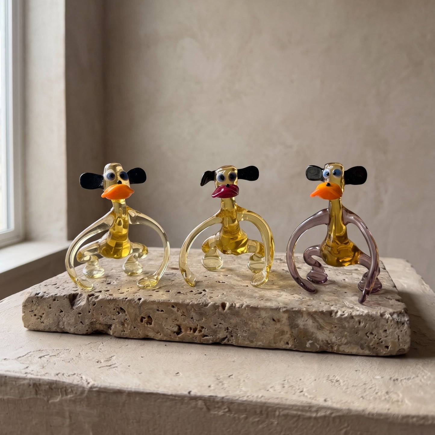 Three Lanky Ape lampworked glass monkey figurines in amber glass with different lip colors seated on a travertine slab, handcrafted by Glass by Iness in L'viv Ukraine
