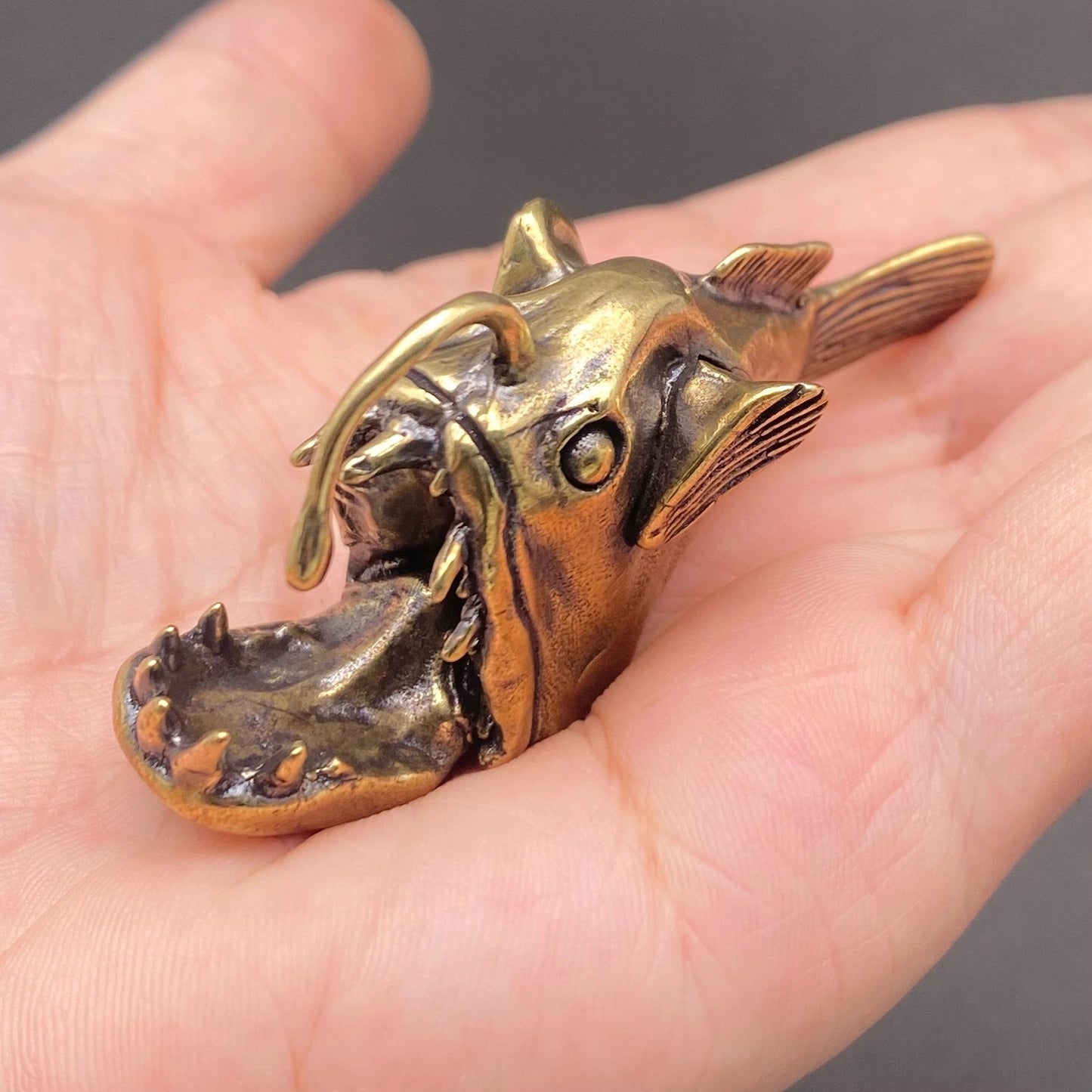 Brass anglerfish figurine resting in an open palm, showing its palm-sized scale, open jaw, and detailed teeth