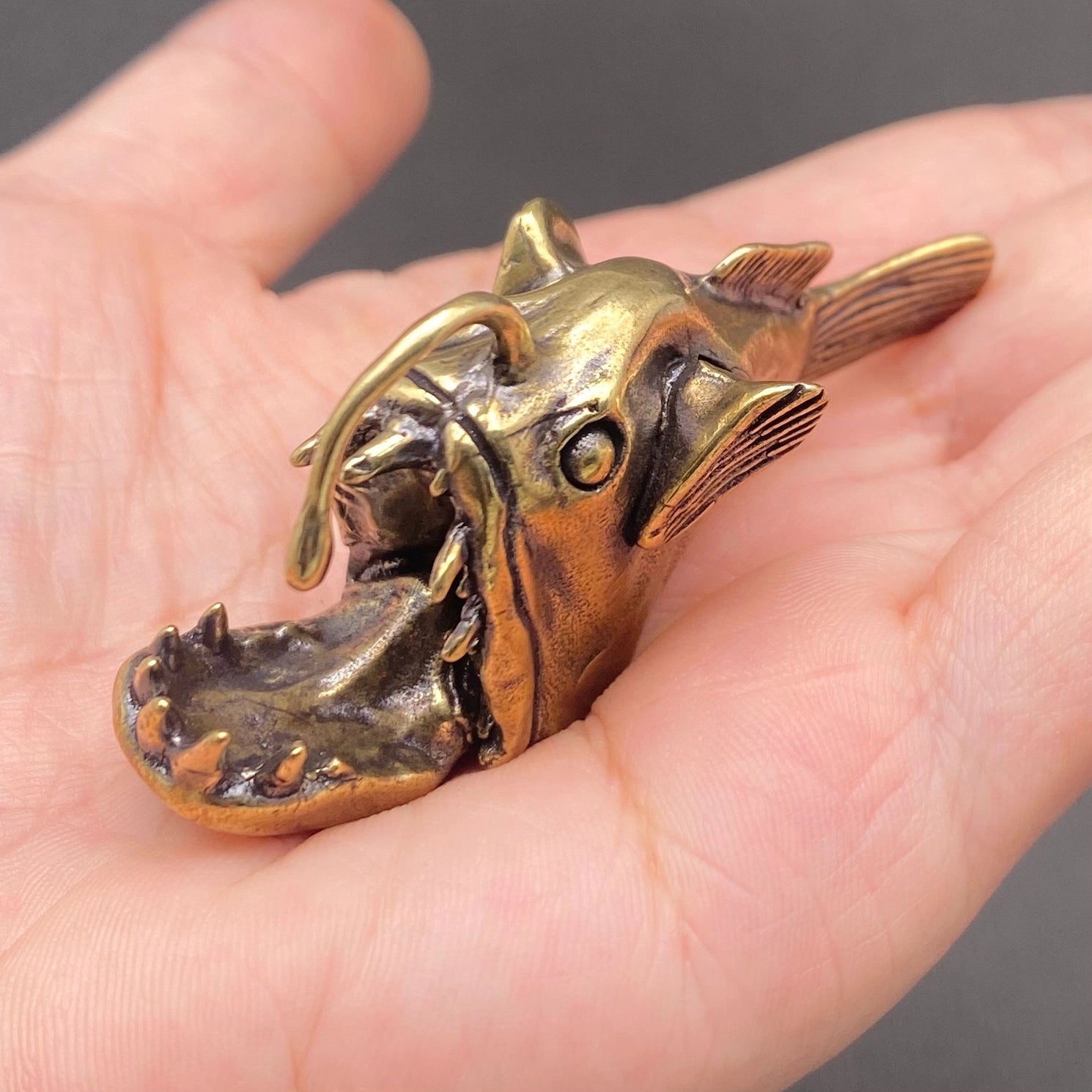 Brass anglerfish figurine resting in an open palm, showing its palm-sized scale, open jaw, and detailed teeth