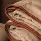 Folded hand-stitched linen quilt in oatmeal with cinnamon terracotta binding, showing layers and hand-stitched dash detail